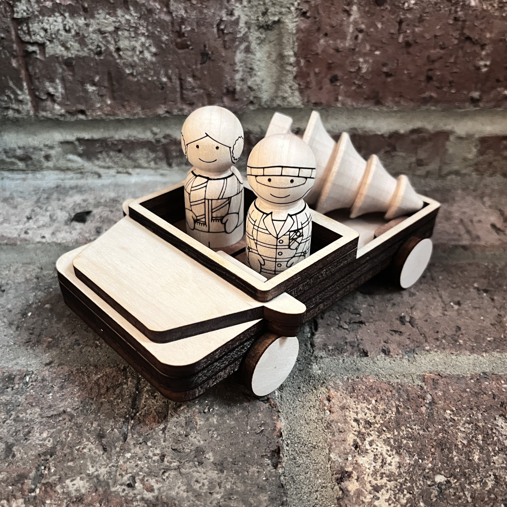 Boy and girl peg doll characters riding in a wooden pickup truck with wooden christmas tree in the truck bed.