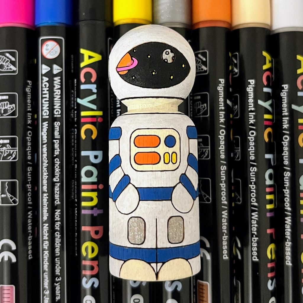 Wooden peg doll laser engraved to look like an astronaut that is colored with acrylic paint markers.